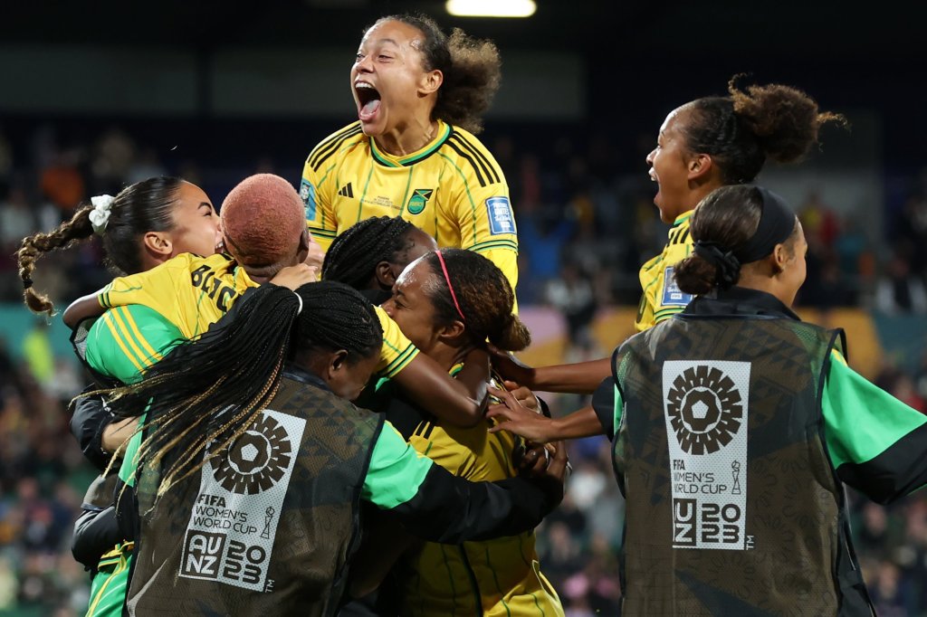 Jogadoras titulares, com camisa amarela com detalhes verdes e pretos, e reservas, de coletes pretos, se abraçam e comemoram gol da Jamaica na Copa do Mundo feminina 2023