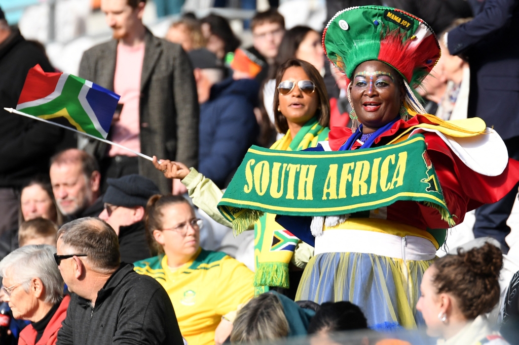 Torcedoras da África do Sul durante a Copa do Mundo feminina. Em primeiro plano, uma mulher negra usa vestes coloridas e segura uma faixa com o nome "South Africa" escrito