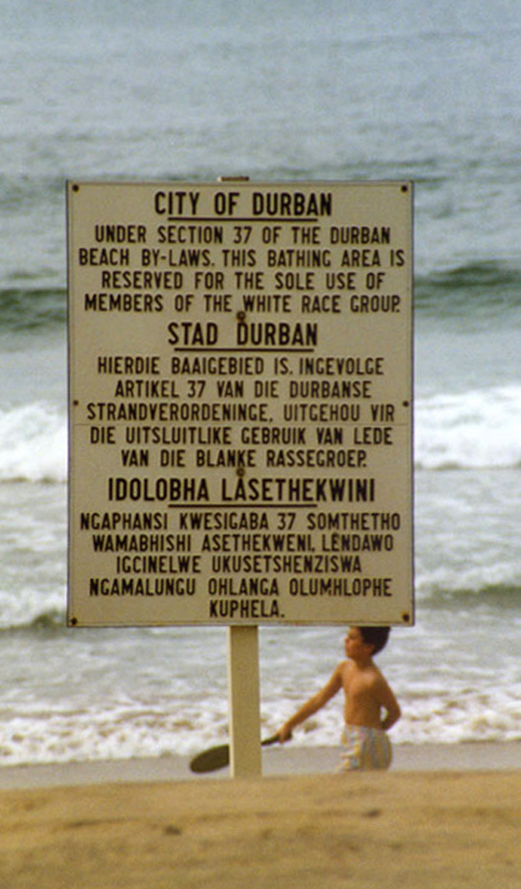 Placa de cor bege com texto em letras pretas fincada na praia de Durban, África do Sul, com o mar e um menino com raquete na mão ao fundo. A mensagem, escrita em inglês, africâner e zulu, diz que só brancos podiam frequentar o local