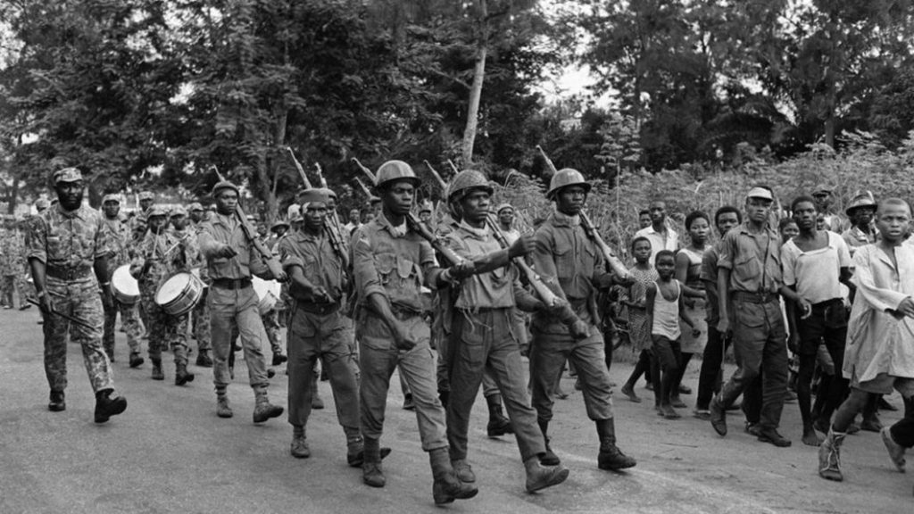 Foto em preto e branco na qual soldados armados marcam em rua da Nigéria. Adultos e crianças observam e acompanham a marcha ao lado dos soldados